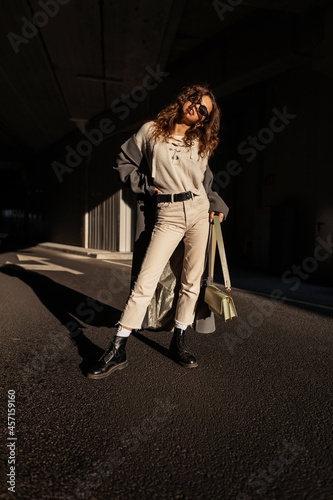 Elegant pretty girl with curly hair and sunglasses in fashionable clothes look: long coat, sweater, pants, shoes and a handbag stands in the city in the sunlight and shade. Urban female style