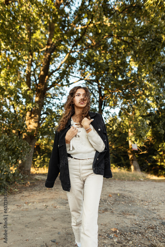 Stylish beautiful young curly-haired model girl in a fashionable black denim jacket and sweater walks in the park Stylish beautiful young curly-haired model girl in a fashionable black denim jacket and sweater walks in the park