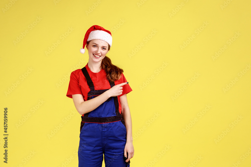 Handy woman looks at camera with smile, pointing finger aside, showing copy space for advertisement, wearing blue overalls and santa claus hat. Indoor studio shot isolated on yellow background. Handy woman looks at camera with smile, pointing finger aside, showing copy space for advertisement, wearing blue overalls and santa claus hat. Indoor studio shot isolated on yellow background.