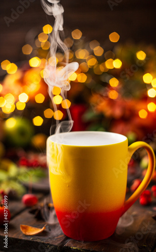 a red and yellow mug with a steaming drink. Fruits and spices are all around on a wooden table. Yellow lights of garlands are burning behind
