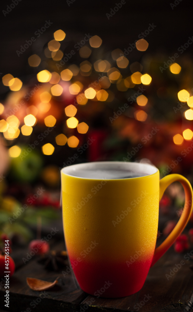 Fototapeta premium a red and yellow mug with a steaming drink. Fruits and spices are all around on a wooden table. Yellow lights of garlands are burning behind