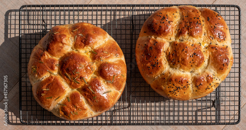 Two traditional home cooked Jewish Challah bread loaves, on a wire tray. One is topped with nigella seeds and the other with rosemary.  photo