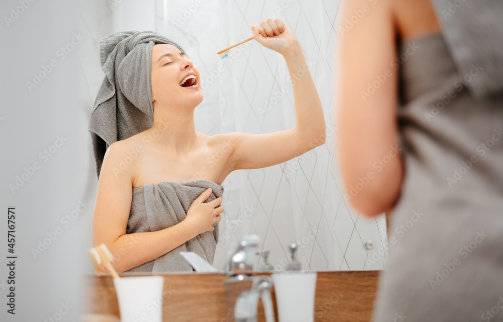 Fototapeta premium Young woman with a towel on her head brushes her teeth and sings as if the toothbrush is a microphone. Concept of dentistry and care.