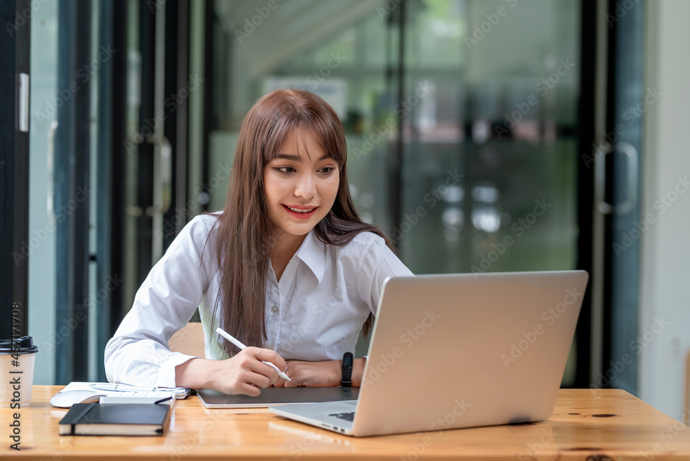 Young Asian businesswoman working holding a pen using a taking note tablet looking at the laptop screen at the office. Young Asian businesswoman working holding a pen using a taking note tablet looking at the laptop screen at the office.