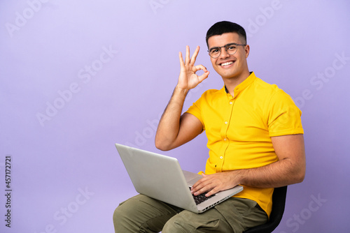 Young man sitting on a chair with laptop showing ok sign with fingers © luismolinero