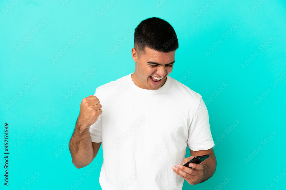 Young handsome man over isolated blue background using mobile phone and doing victory gesture Young handsome man over isolated blue background using mobile phone and doing victory gesture