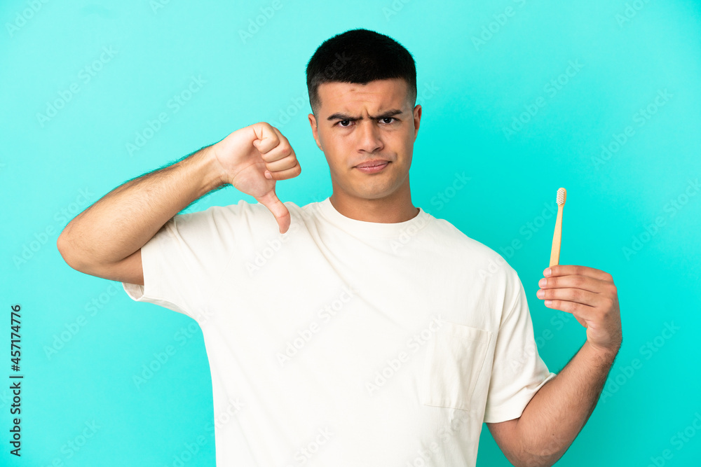Young handsome man brushing teeth over isolated blue background showing thumb down with negative expression