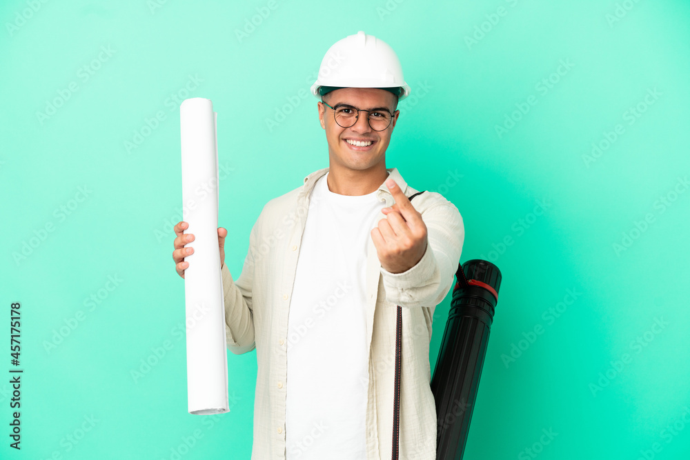 Young architect man holding blueprints over isolated background doing coming gesture Young architect man holding blueprints over isolated background doing coming gesture