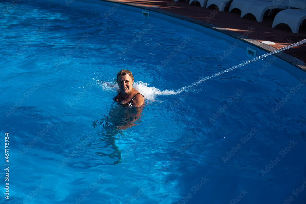 An adult, smiling woman enjoys a hydro massage of the back and neck with a jet of water in the pool at the spa on a trip. 