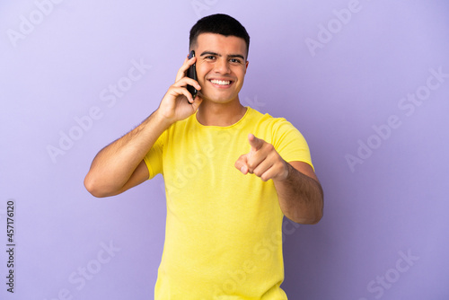 Young handsome man using mobile phone over isolated purple background surprised and pointing front