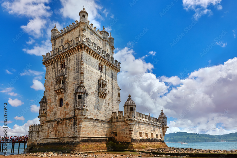 Lisbon, Portugal. Tower Belem at coast of river Tagus. Stones and mussels during low tide. Sunny summer day with blue sky and clouds. Embankment alley with palm tree leaves.