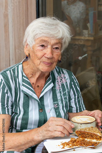 An old, elderly woman is having breakfast, eating cake and drinking coffee at a table in a summer cafe on the street. High quality photo