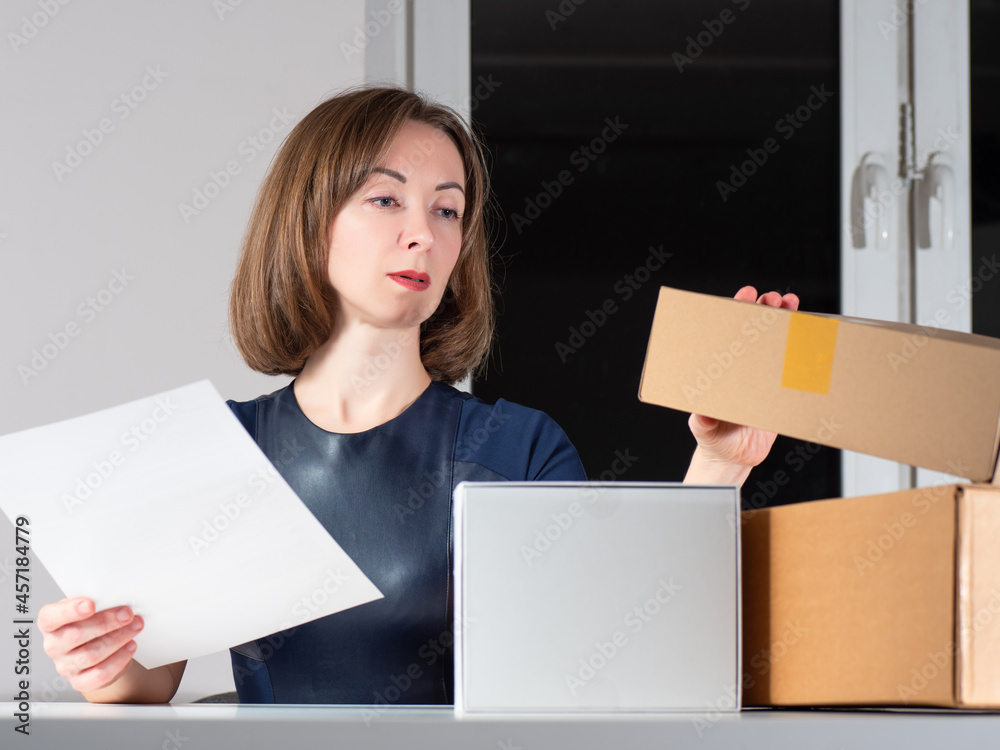 Woman next to cardboard boxes. She closes he box. Businesswoman holds a sheet of paper in her hand. Concept - she is engaged in home business. Woman home business owner. Girl on background of window Woman next to cardboard boxes. She closes he box. Businesswoman holds a sheet of paper in her hand. Concept - she is engaged in home business. Woman home business owner. Girl on background of window