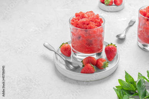 Strawberry granita with mint sicilian frozen summer dessert in glass on a white plate on light grey stone background with copy space. Horizontal orientation. Selective focus.