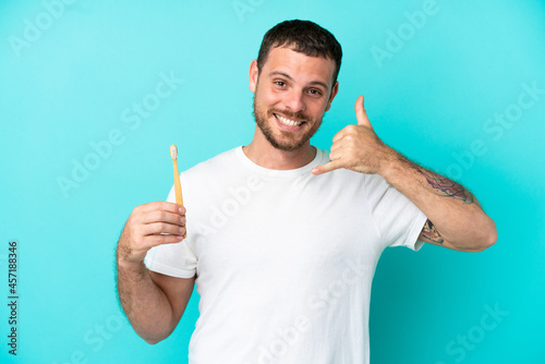 Young Brazilian man brushing teeth isolated on blue background making phone gesture. Call me back sign