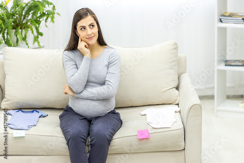 Photo of pregnant woman sitting on the sofa and childish clothes near.