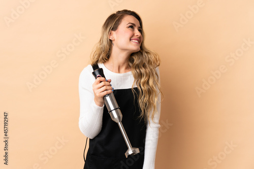 Young brazilian woman using hand blender isolated on beige background thinking an idea while looking up