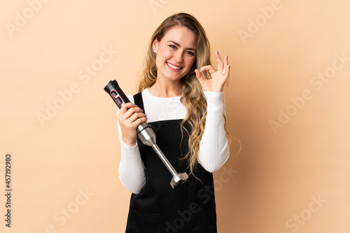 Young brazilian woman using hand blender isolated on beige background showing ok sign with fingers