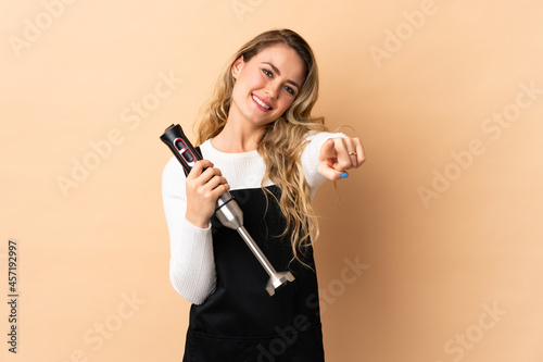 Young brazilian woman using hand blender isolated on beige background pointing front with happy expression