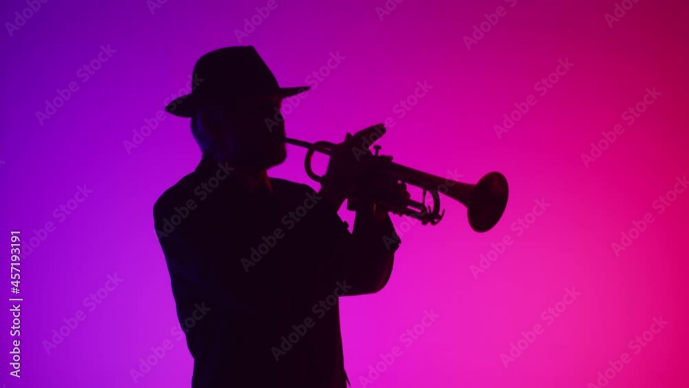 Man in jacket and hat plays music on trumpet in room lit by purple neon, front view. Jazz musician at suit plays trumpet