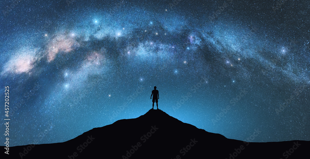 Milky Way arch and man on the mountain peak at starry night. Silhouette of alone guy, blue sky with bright stars in summer. Galaxy. Space background. Landscape with arched milky way. Travel and nature Milky Way arch and man on the mountain peak at starry night. Silhouette of alone guy, blue sky with bright stars in summer. Galaxy. Space background. Landscape with arched milky way. Travel and nature