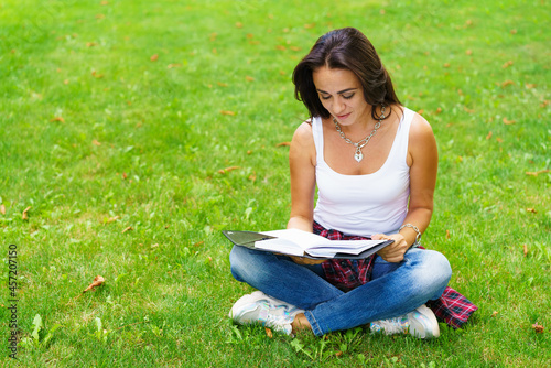 Smiling female student sitting on grass with notepad in hand, preparing for exams. Education and remote work concept. Soft selective focus. Caucasian girl with a pensive look © Екатерина Переславце