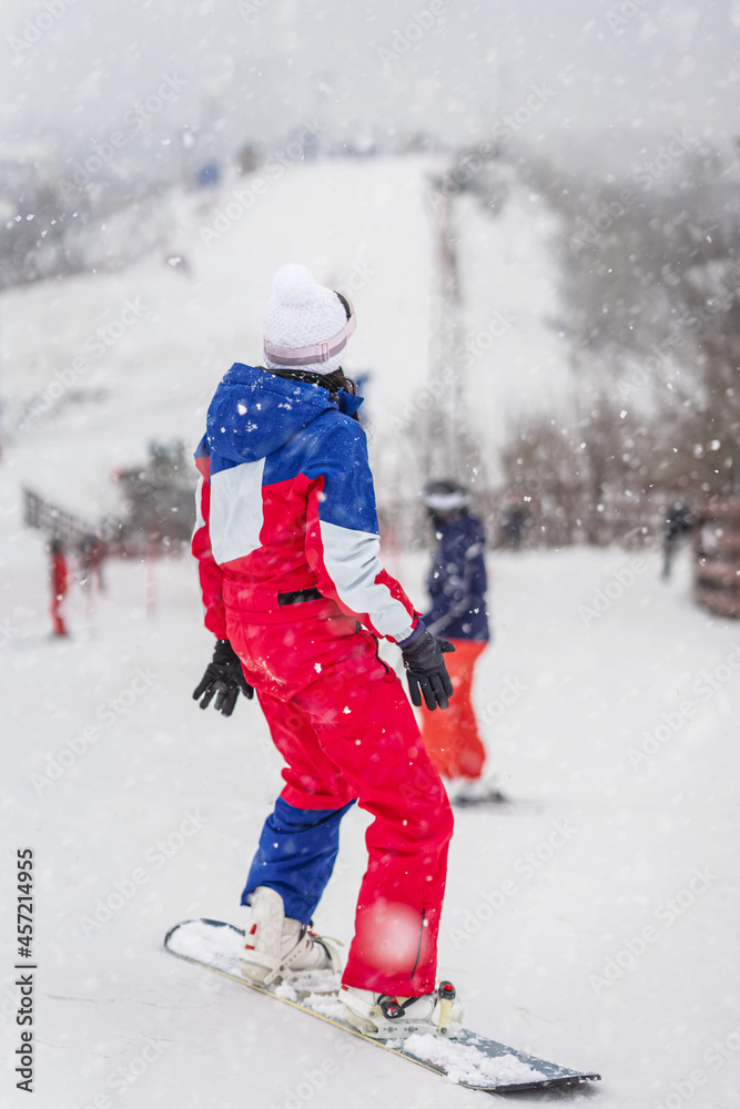 Back view close-up of girl snowboarder standing on ski slope in bright tracksuit. Winter holiday, outdoor sport