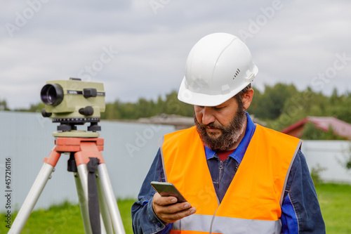 A civil engineer with an optical level looks into a mobile phone. A bearded man is looking for information on his mobile phone.