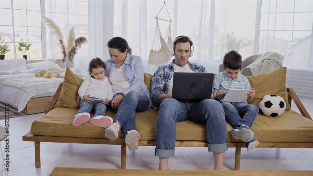 Addicted to gadgets parents and kids busy on laptop, tablet and cellphone while sitting on sofa in living room. Inactive family with preadolescent children spending joint leisure time using gadgets