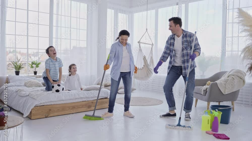 Cheerful parents dancing fun with mop and sweeping brush while their little children jumping on bed and laughing. Happy family with kids having rest while doing household chores together Cheerful parents dancing fun with mop and sweeping brush while their little children jumping on bed and laughing. Happy family with kids having rest while doing household chores together