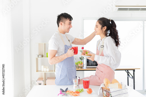 asian man and asian woman eating meal in the morning, they have breakfast in kitchen room, he holding hot coffee cup, she holding sandwich with hand, they feeling happy and smile, happiness honeymoon 