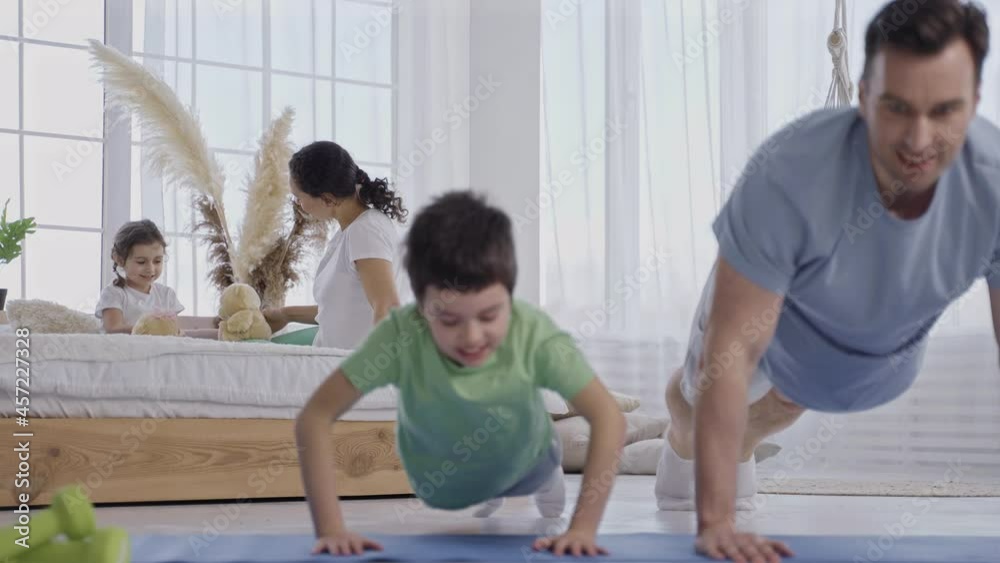 Athletic father doing push ups together with preadolescent boy blurry on foreground while mom and 3-years-old girl playing with teddy bears on bed. Friendly family during gaming and sports activities Athletic father doing push ups together with preadolescent boy blurry on foreground while mom and 3-years-old girl playing with teddy bears on bed. Friendly family during gaming and sports activities