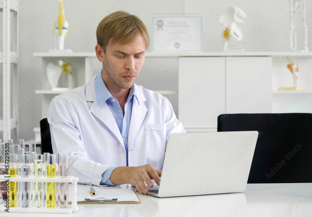 Young caucasian scientist use computers to research chemical compounds on the internet. Working atmosphere in chemical laboratory. Test tubes filled with chemicals on the table. Young caucasian scientist use computers to research chemical compounds on the internet. Working atmosphere in chemical laboratory. Test tubes filled with chemicals on the table.
