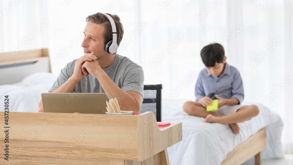 Young caucasian father working with his laptop and wearing headphones at home while his kid playing in the background. Concept work from home