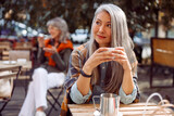 Senior cafe guests, focus on dreamful long haired mature lady holding cup of drink at table on outdoors cafe terrace on autumn day