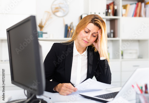 Worried young woman manager working at laptop with documents in modern office