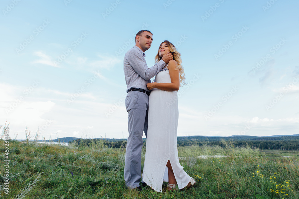 Happy young couple, expecting a baby, walking in summer rainy day on a field. Man hugs his pretty pregnant woman in a white dress. Waiting for the baby, pregnancy concept. Happy young couple, expecting a baby, walking in summer rainy day on a field. Man hugs his pretty pregnant woman in a white dress. Waiting for the baby, pregnancy concept.