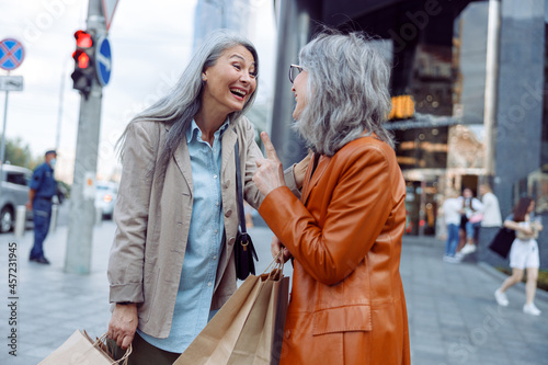 Cheerful mature women in stylish outfit with shopping bags talk on modern city street on autumn day. Friends spend time together