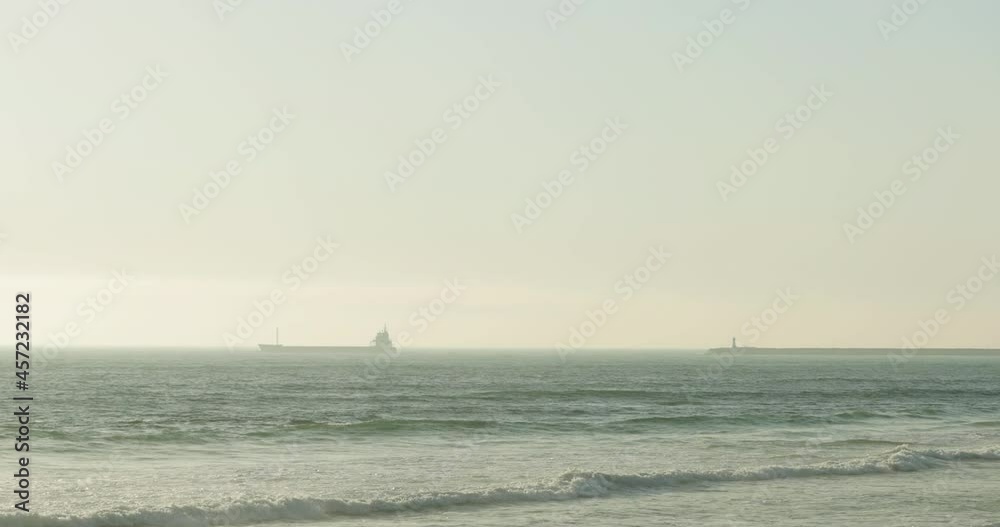 Cargo Ship Cruising On A Foggy Ocean With Waves Coming To The Beach Shore In Figueira da Foz, Portugal. - wide shot