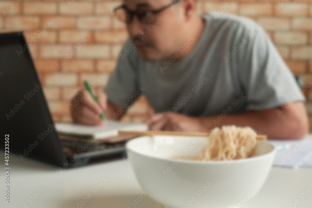 Blur focus shot, Thai male worker busy working with laptop, use chopsticks to eat instant noodles during office lunch breaks, because quick and tasty. Over time Asian fast food, unhealthy lifestyle. Blur focus shot, Thai male worker busy working with laptop, use chopsticks to eat instant noodles during office lunch breaks, because quick and tasty. Over time Asian fast food, unhealthy lifestyle.