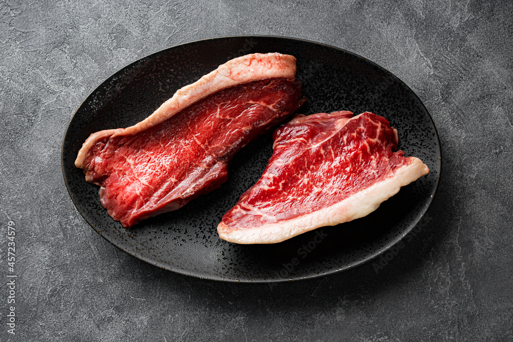  Raw marbled beef steaks on the black plate and dark gray background, top view. Raw beef meat on the kitchen table