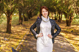 Portrait of a young beautiful brunette girl in a black leather jacket and a knitted sweater with short hair smiling while standing on the road in an autumn park. Warm autumn day