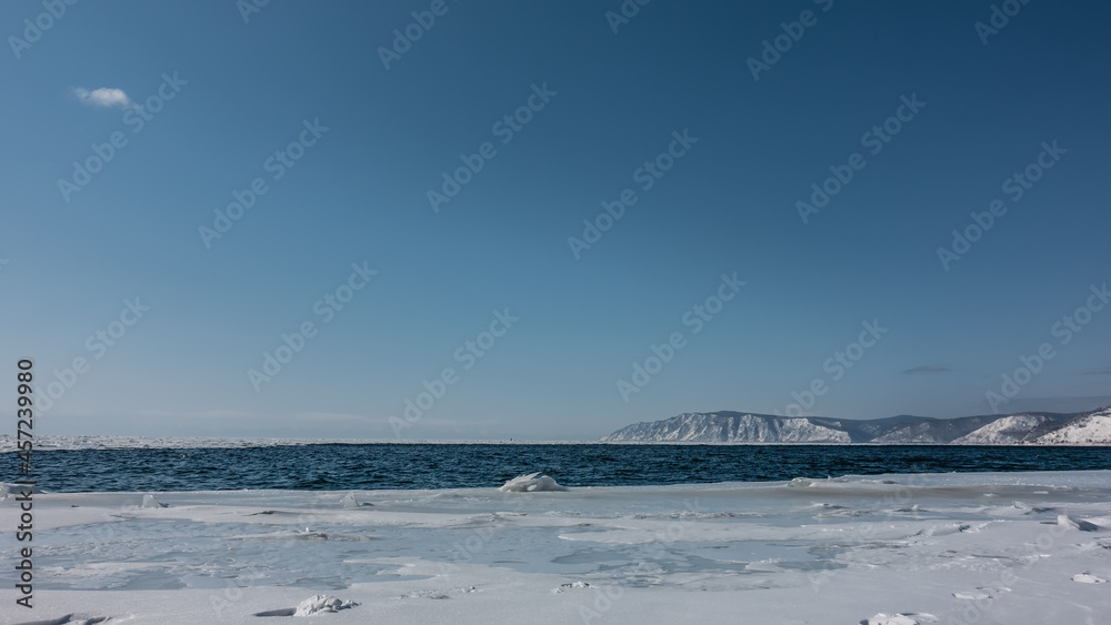 Fototapeta premium Snowdrifts lie on the bank of a non-freezing river. Ripples on the blue water. A mountain range on the background of an azure sky. Angara