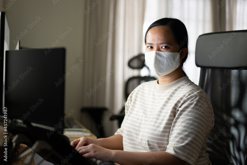 Asian woman with protection mask is working at home looking at camera
Asian woman with protection mask is working at home looking at camera