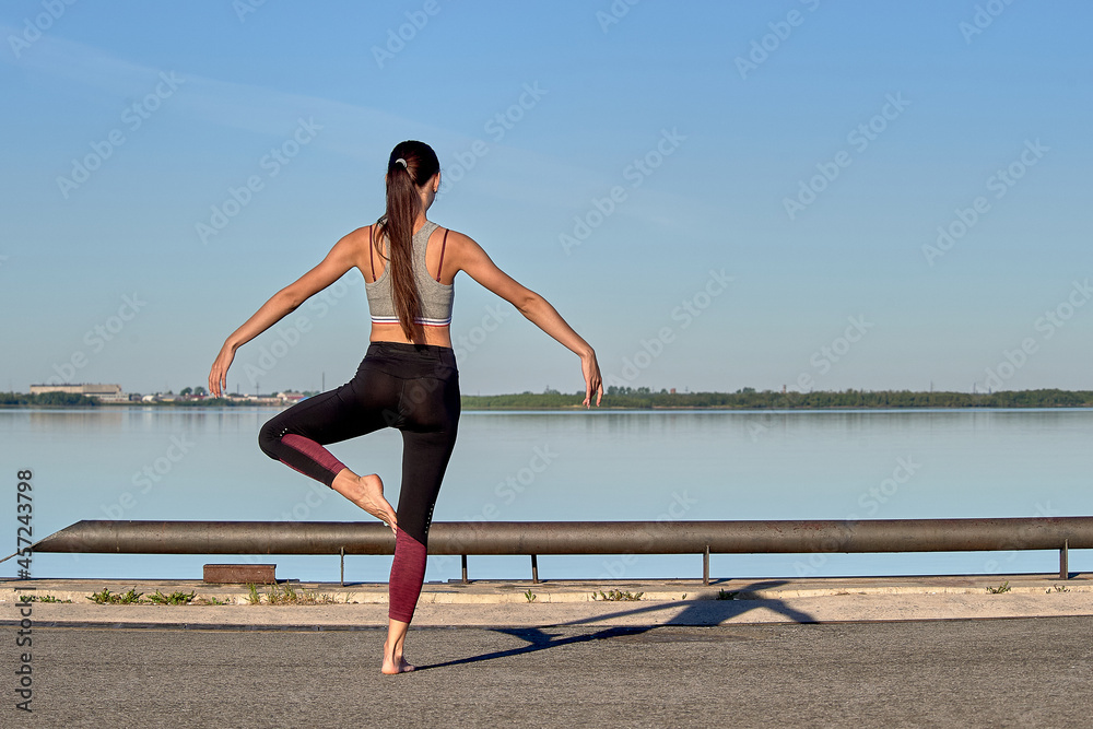 A young woman does a warm-up on the pier in the early morning, against the background of the river