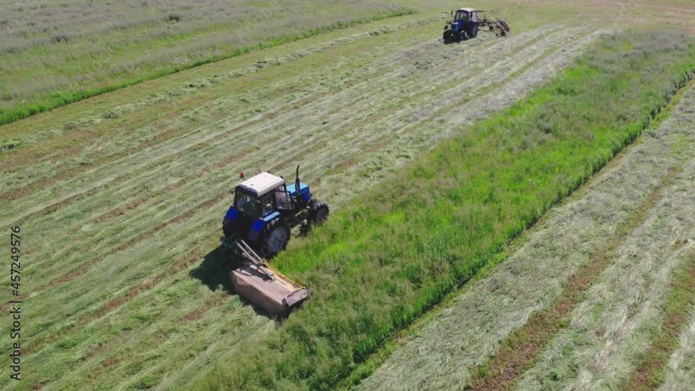 Aerial view of two tractors mowing and raking grass in the field in summer. Agricultural machines moving towards each other. Concept of seasonal hay harvesting Aerial view of two tractors mowing and raking grass in the field in summer. Agricultural machines moving towards each other. Concept of seasonal hay harvesting