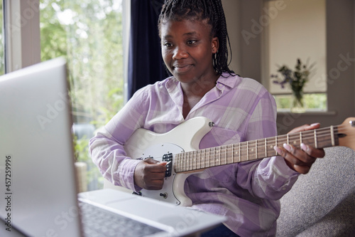 Teenage Girl At Home Learning To Play Electric Guitar With Online Lesson On Laptop Computer