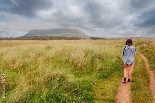 Young slim teenager girl walking on a small road towards Strandhill town, Knocknarea hill in cloudy sky in the background. County Sligo, Ireland. Irish landscape. Popular landmark. © mark_gusev