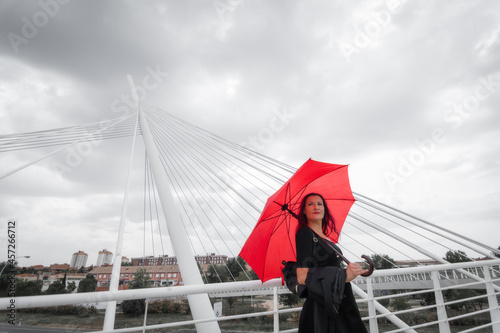 White female business executive smiling with business briefcase, 40s long reddish hair brunette with black dress, wellies and red umbrella looking at camera over modern city bridge. Toledo, Spain