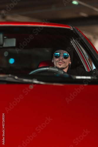 Stylish young man in sunglasses and a hat sits at the wheel of an old red car. male taxi driver driving